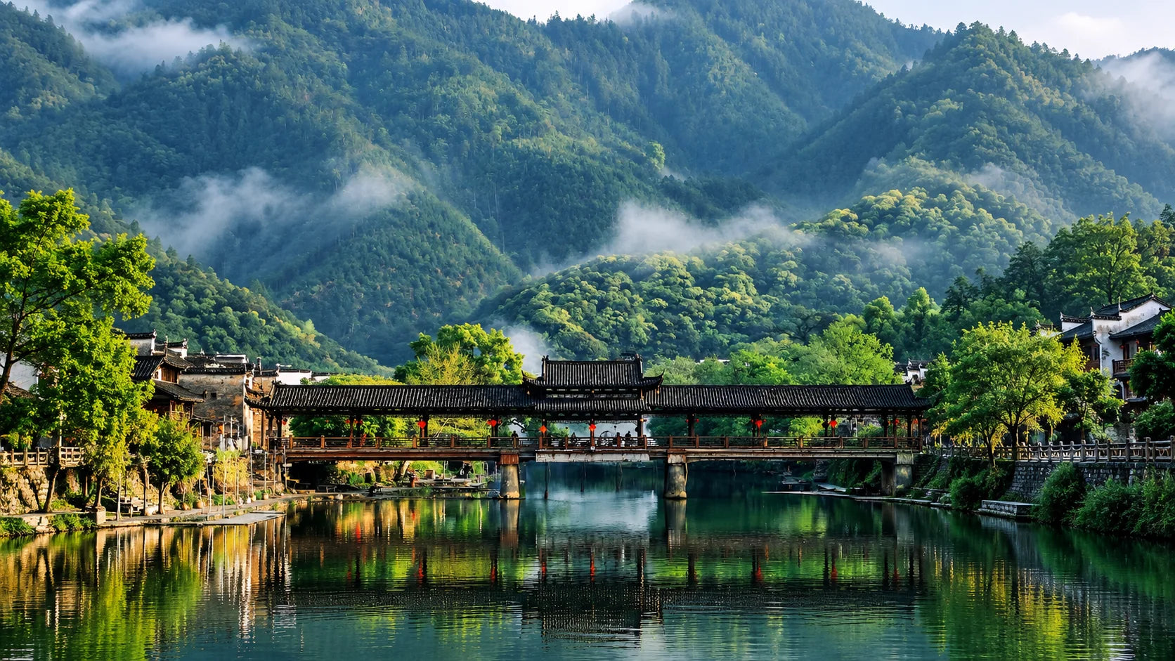 A covered wooden bridge over a still river, with mist rising through forested mountains beyond