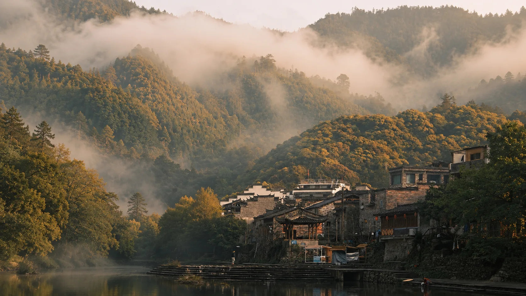 A riverside village below forested hills at dawn, with mist drifting through the valley